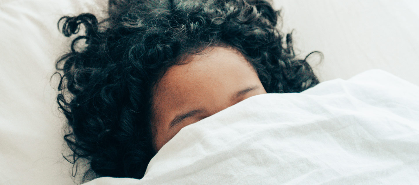 Fille aux cheveux noirs bouclés endormie sur le dos sur le lit recouverte d'une couette jusqu'aux yeux.