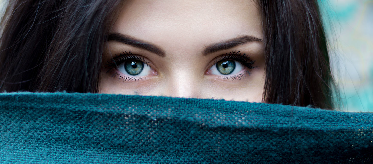 Fille aux longs cheveux noirs et aux yeux verts couverte d'un foulard jusqu'au nez regardant droit devant elle.