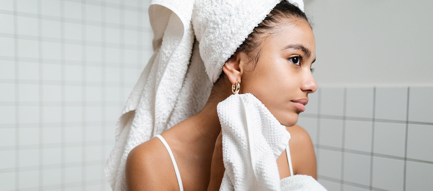 Fille brune sortie de la douche s’essuyant le visage avec une serviette, et avec une autre serviette roulée sur sa tête dans la salle de bain.