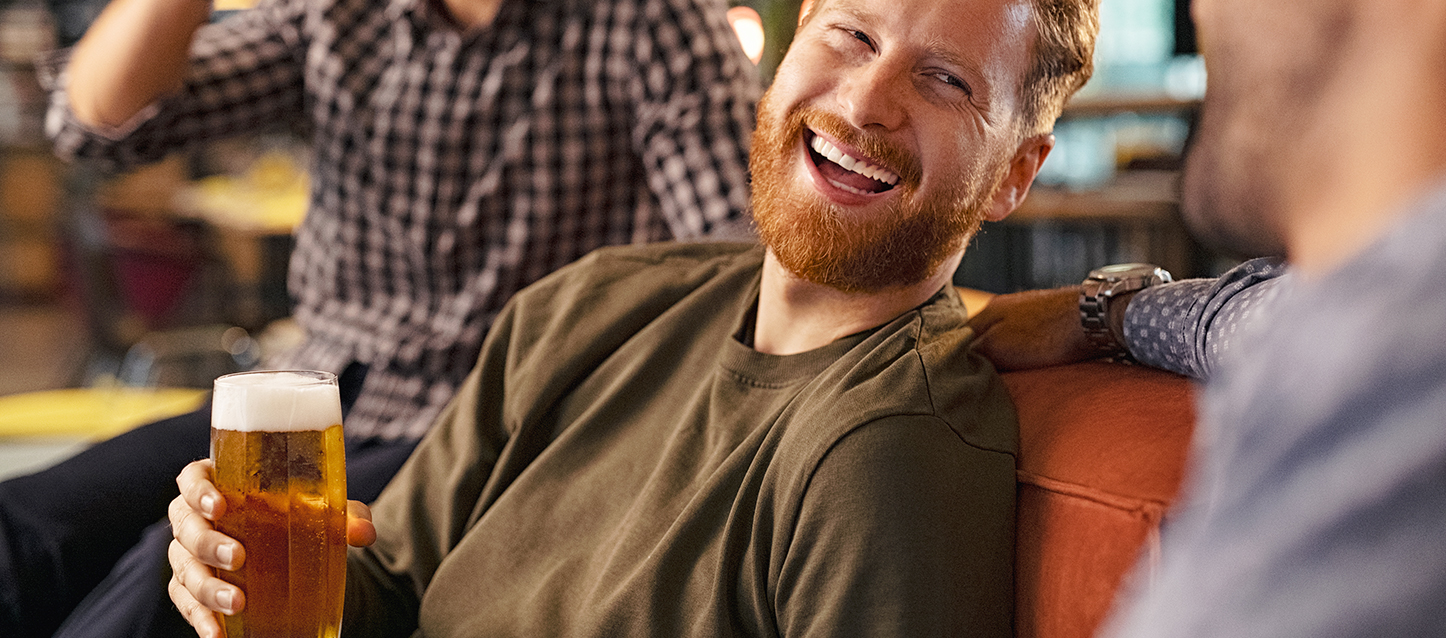 Un gars aux cheveux roux ayant une barbe en riant tenant une bière dans un pub.