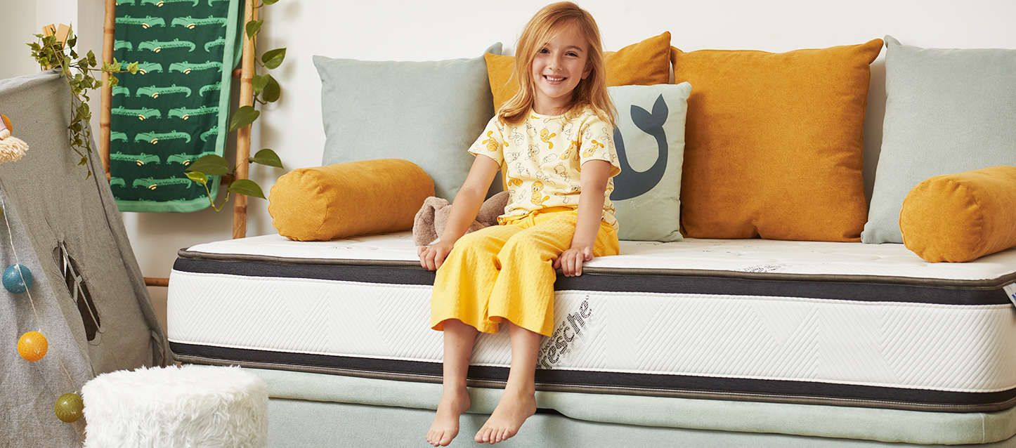 Petite fille aux cheveux blonds souriant dans une chambre assise sur le matelas d’un lit coffre.
