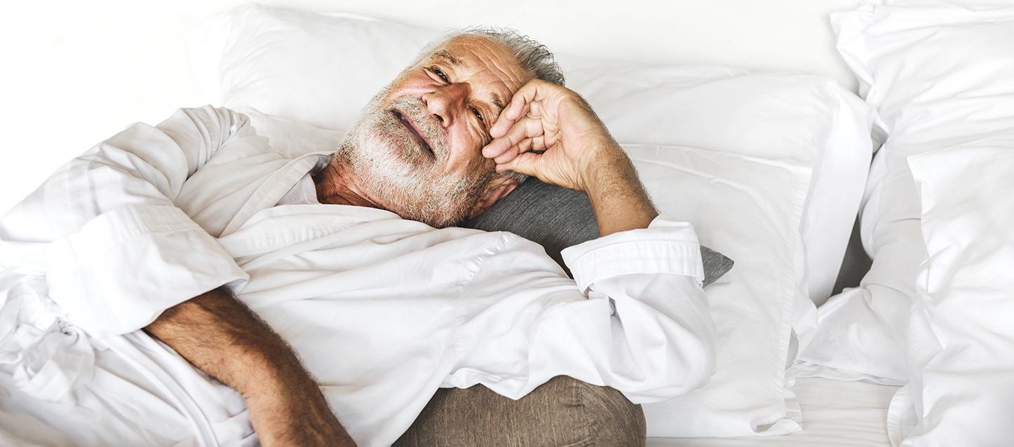 Homme âgé à la barbe blanche allongé sur un lit, sur le dos et sur un coussin gris.