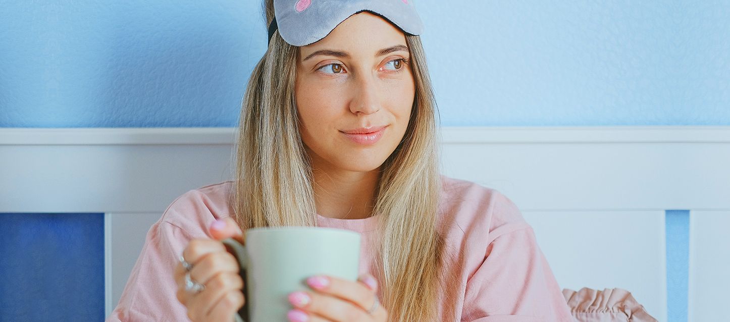 chica rubia de pelo largo con pijama rosa y antifaz gris sujetando una taza verde pastel con ambas manos