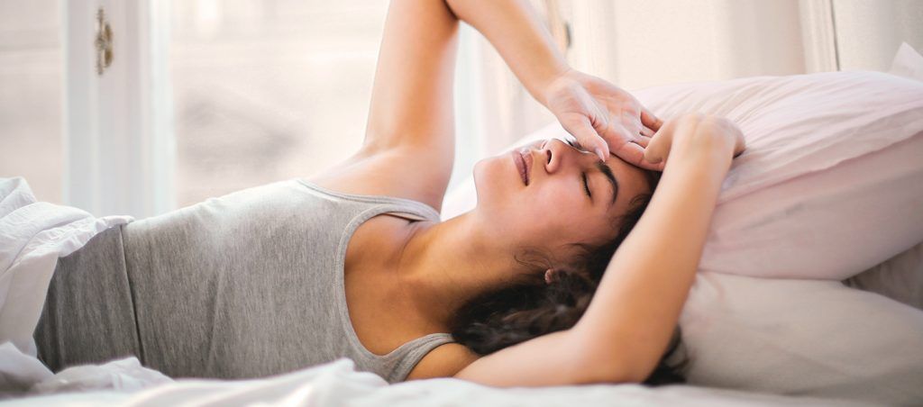 Consecuencias de dormir en un mal colchón chica de media melena morena tapada dentro de la cama tapada con edredon hasta la cintura boca arriba con las manos en la frente con gesto de malestar por haber dormido en un mal colchon
