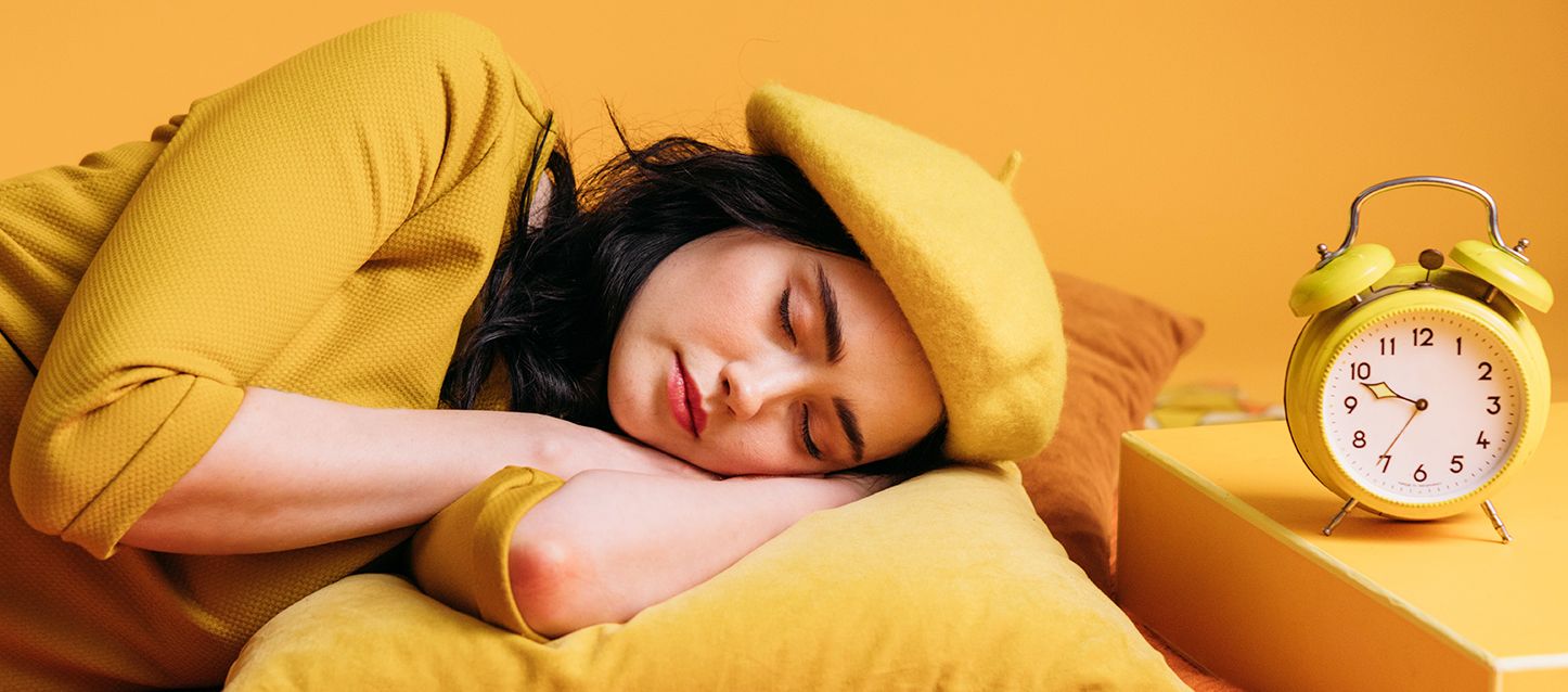 Chica morena de pelo largo negro vestida con vestido y boina amarilla durmiendo de lado apoyada sobre su brazo junto a un despertador amarillo
