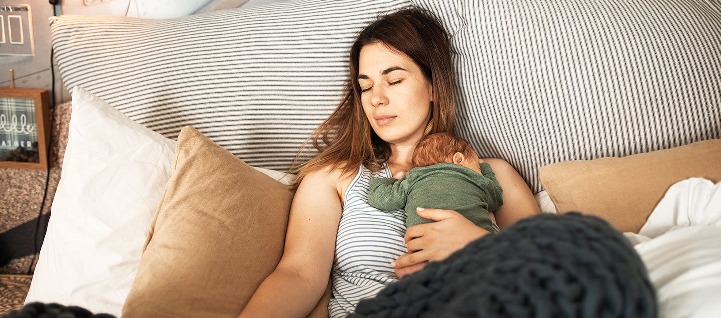 mujer de pelo largo castaño con camiseta a rayas durmiendo en la cama con su bebe encima con pijamita verde
