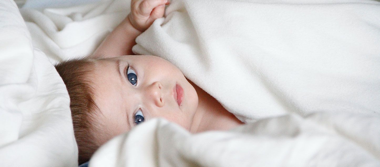 Los niños que no duermen pueden enfermar más niño de pelo castaño con ojos azules tapado con una mantita blanca mirando