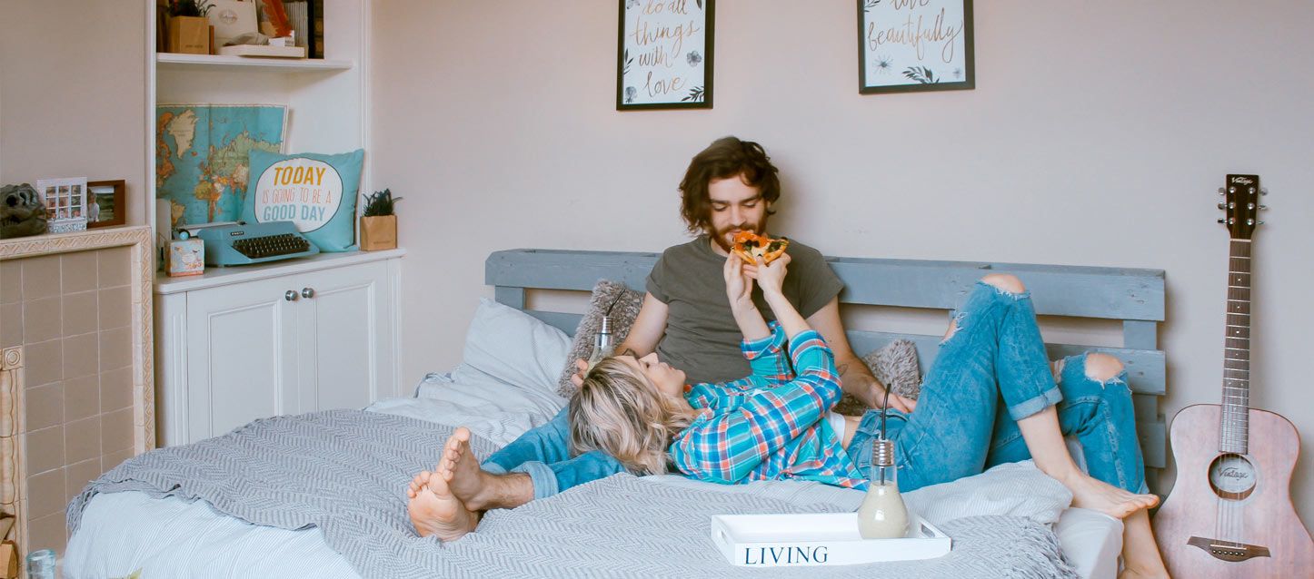 matrimonio joven en un dormitorio con cabecero hecho con palets donde ella esta tumbada sobre las piernas de el y ella le da a comer un trozo de pizza