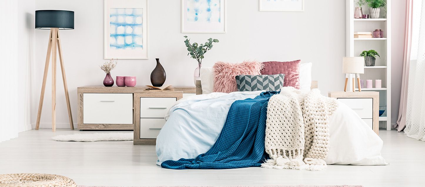 dormitorio fresco con edredon azul, manta de punto, cojines rosas, mesita de noche, lampara de pie y estanteria