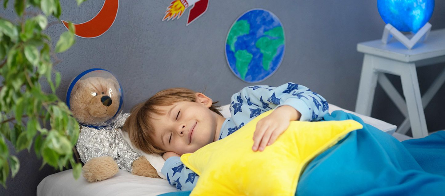 niño pequeño durmiendo boca arriba sonriendo con pijama azul claro con estrellas azul oscuro tapado con una manta azul y sujetando un cojín con forma de estrella amarilla