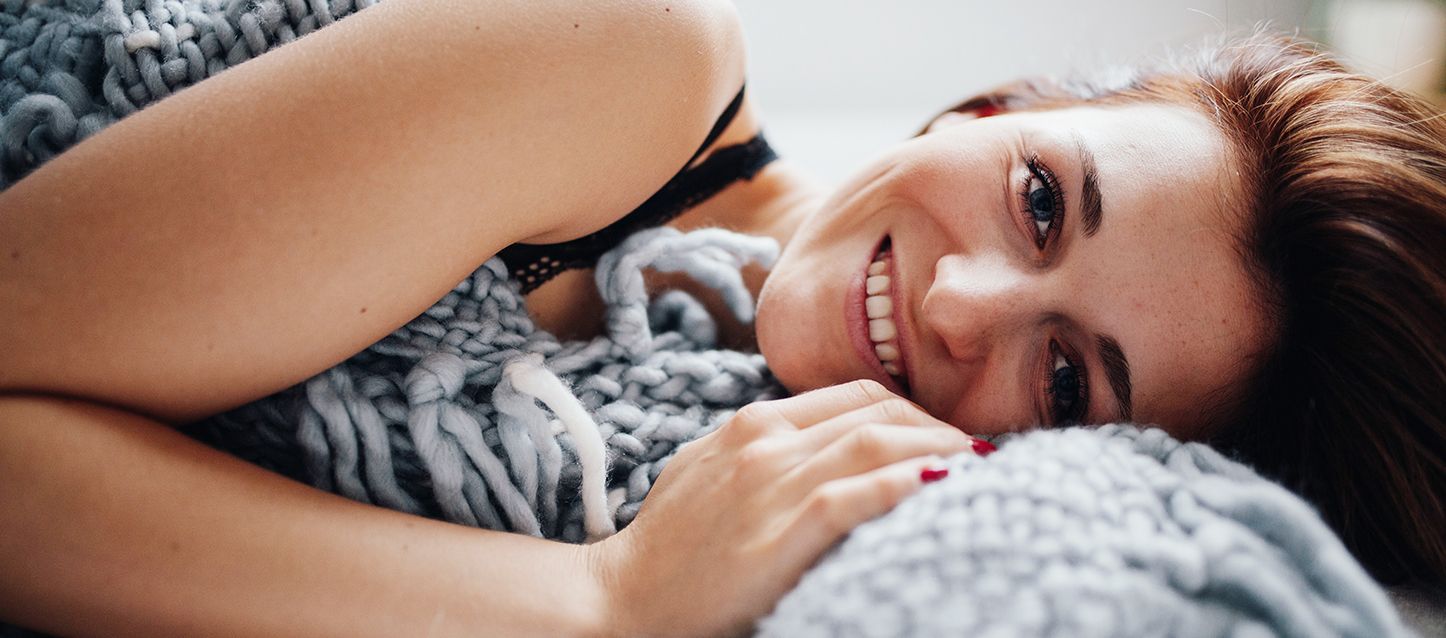 Mujer de pelo largo castaño tumbada en la cama de lado sonriendo y tapada con una manta de punto