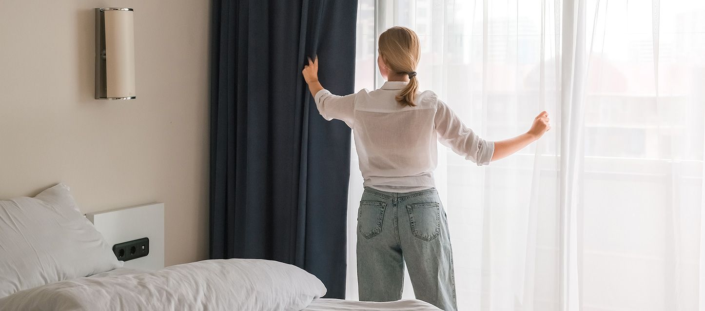 Mujer cogiendo cortinas en una habitación con una cama y una lampara de pared