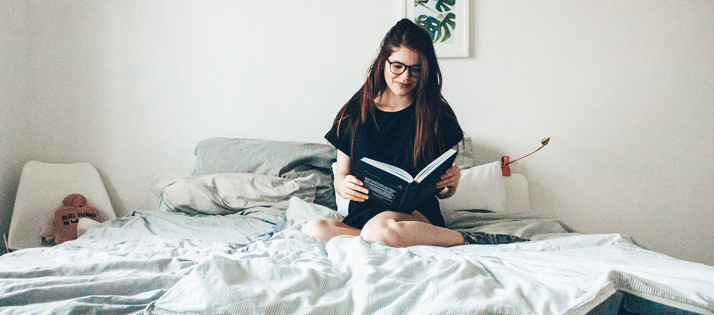 rapariga de cabelo comprido sentada na cama a ler um livro, e vestida com camiseta preta