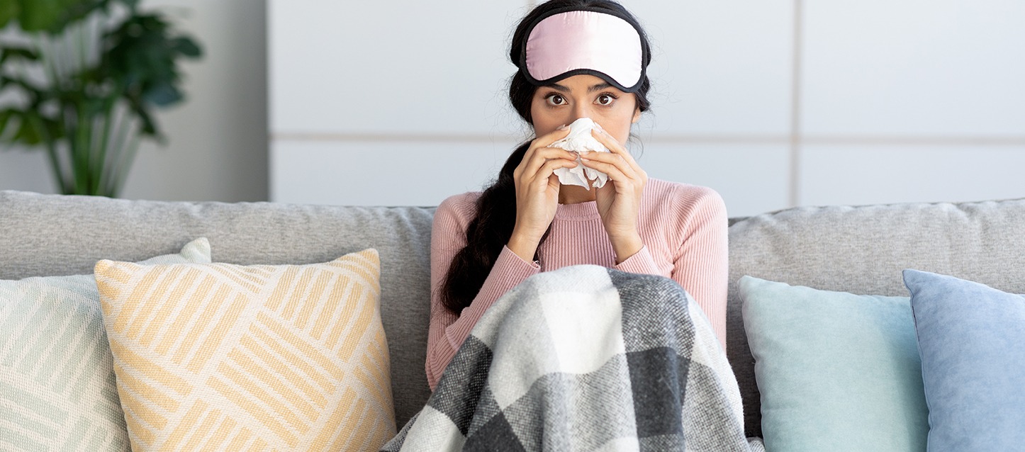 rapariga morena de cabelo comprido, com camisola de cor rosa, sentada no sofá, tapada com uma manta aos quadrados, com um lenço no nariz por sofrer de alergias
