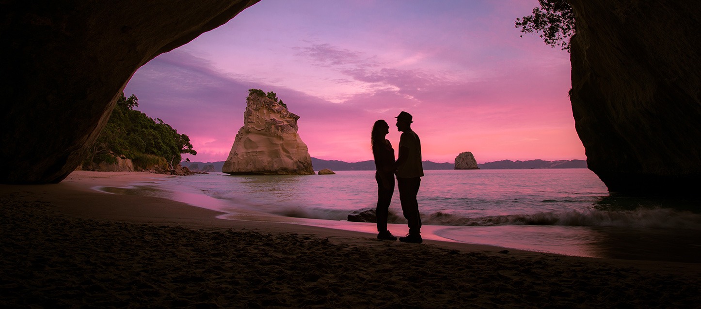 casal de pé numa praia paradisíaca, olhando-se um ao outro enquanto amanhece