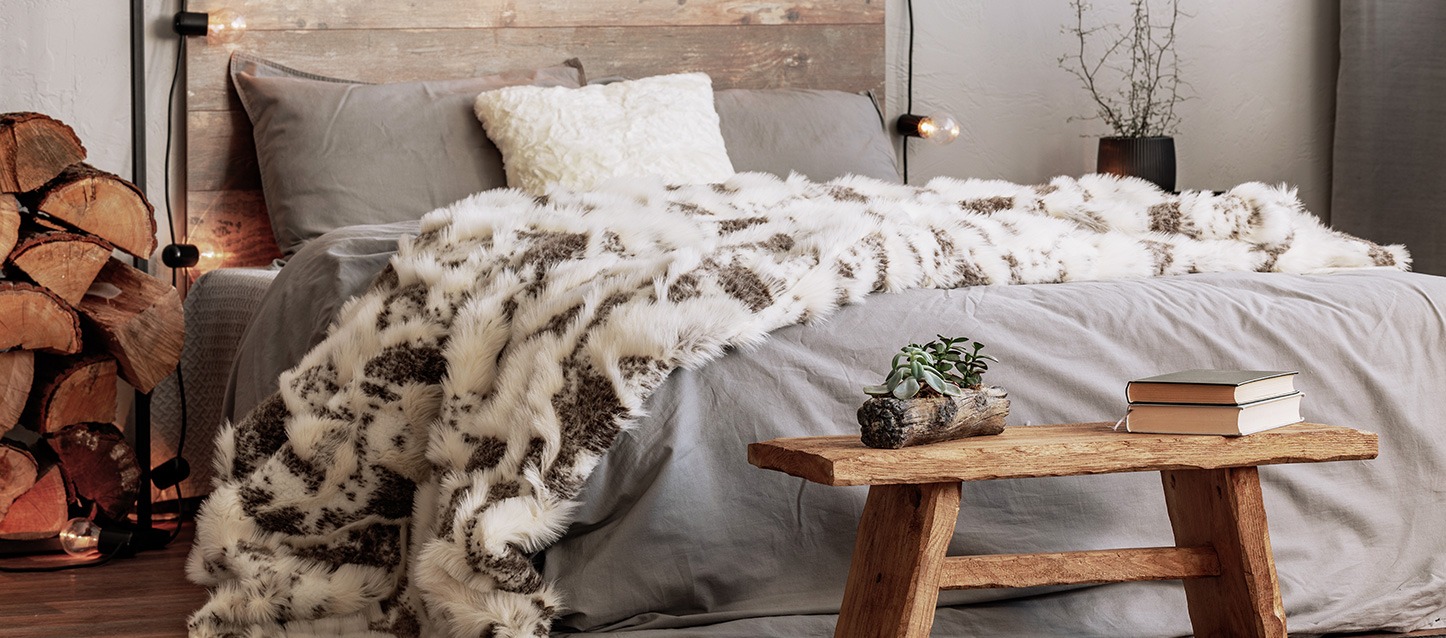quarto com cama e edredão cinza, manta de inverno, travesseiro, duas almofadas e mesinha de madeira com planta e dois livros