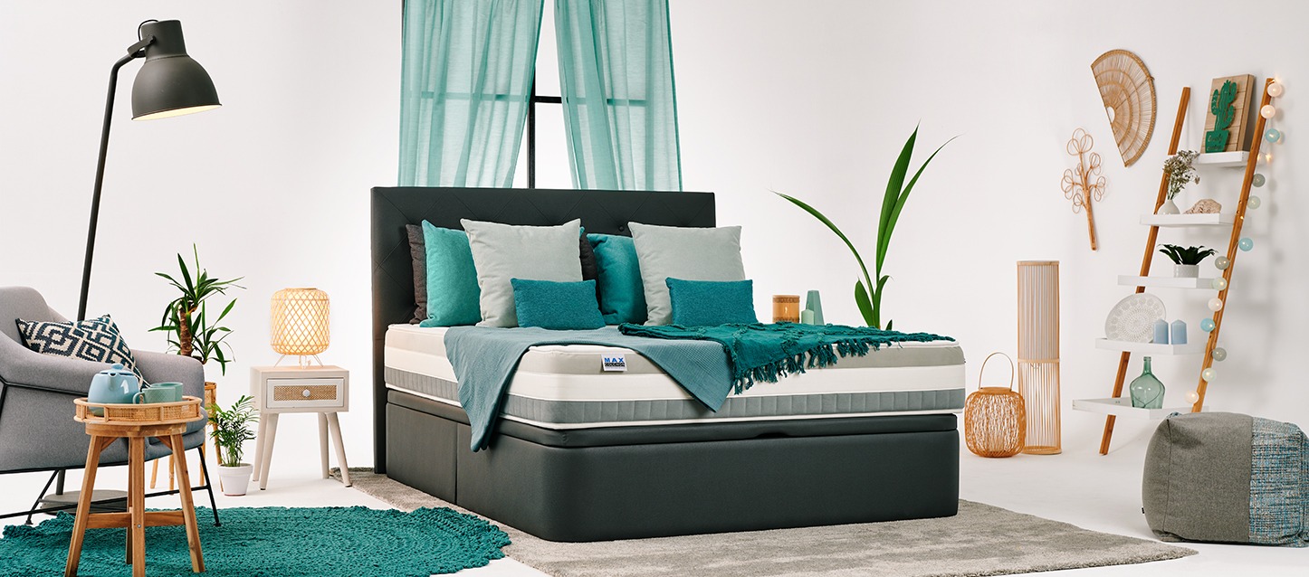 quarto com sommier revestido, colchão, colcha, 8 travesseiros de diferentes tamanhos, mesa, cadeira e estantes com objetos decorativos