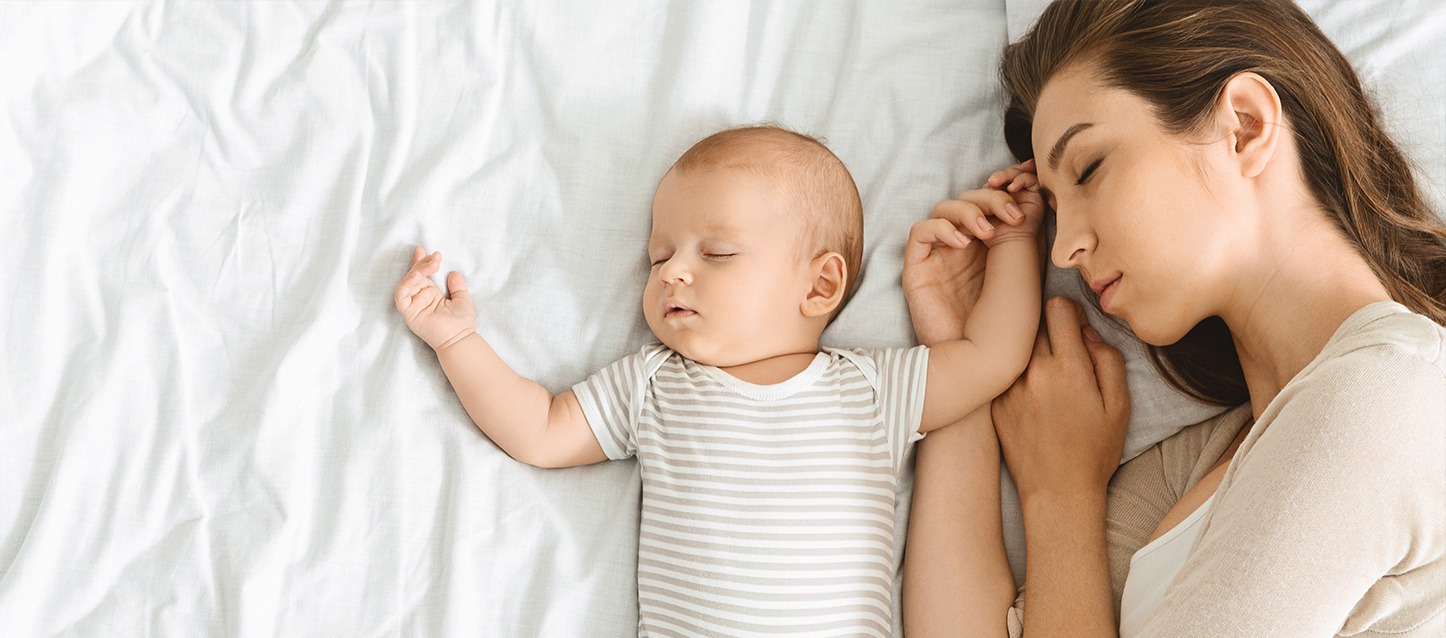 Bebé e a sua mãe a dormirem juntos numa cama de lençóis brancos.