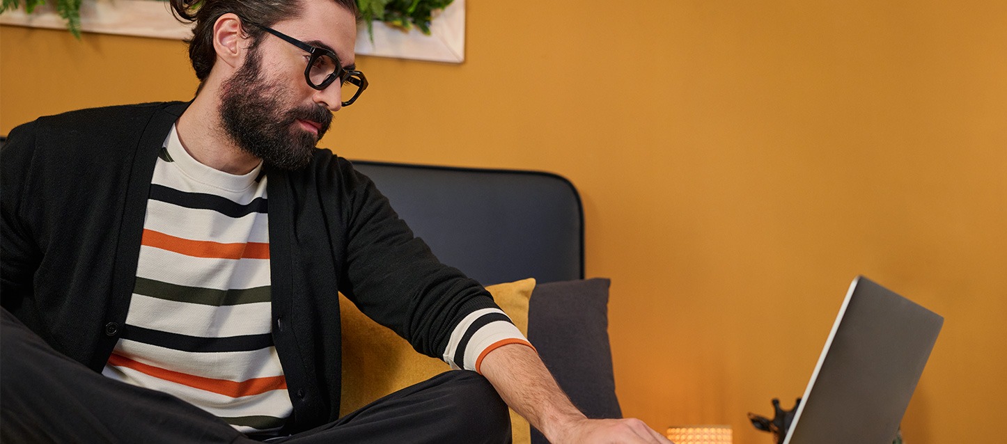 Homem com óculos sentado numa cama a trabalhar com um computador portátil.